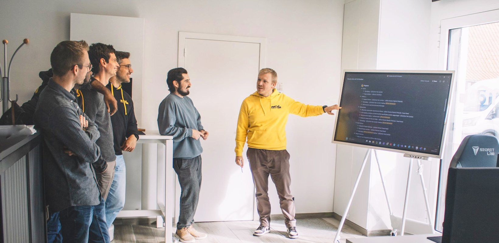 Mad IT developer geeft presentatie aan collega’s over softwarecode op groot scherm, in een agile werkomgeving gericht op samenwerking, kennisdeling en Microsoft-technologie.