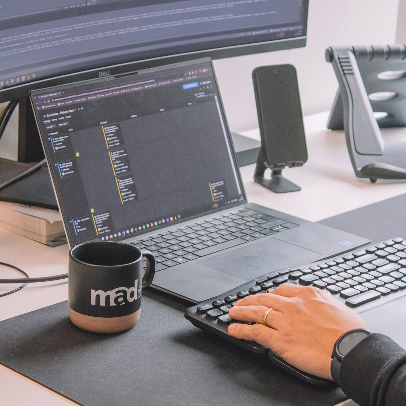 Hand van Mad IT-medewerker op toetsenbord naast laptop met programmeercode op scherm en Mad IT-koffietas — moderne werkplek voor .NET softwareontwikkeling.